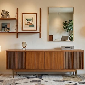 May include: A mid-century modern living room featuring a wooden cabinet, shelves, and a floor lamp. The cabinet has a light brown finish with vertical wood slats on the sides. A framed artwork and a mirror are also visible.