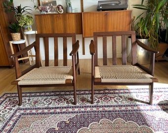 May include: Two mid-century modern chairs with woven cane seats and dark wood frames. The chairs are sitting on a patterned rug in front of a wooden cabinet.