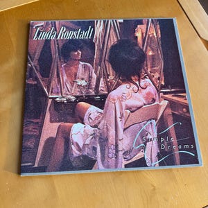 May include: A vintage vinyl record album cover featuring Linda Ronstadt. The cover art shows a woman in a floral robe, seated in front of a mirror. The album title "Simple Dreams" is visible, along with the artist's name.