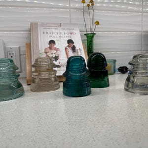 Può includere: Cinque isolatori in vetro vintage, due sono in vetro trasparente e tre sono in vetro verde. Gli isolatori sono disposti in fila su un bancone bianco.