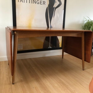 May include: A wooden extendable dining table with a light brown finish. The table has two leaves that can be added to extend the length of the table. The table has a simple, mid-century modern design.