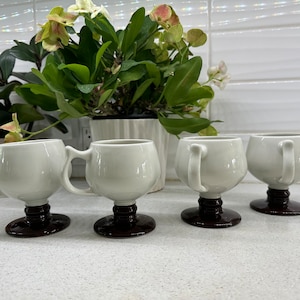 Può includere: Quattro tazze in ceramica bianca con manici e basi marroni. Le tazze sono disposte in fila su un bancone bianco.