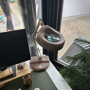 Può includere: Una lampada con lente d'ingrandimento beige con una grande lente rotonda si trova su una scrivania bianca con un tappetino da scrivania verde. Una tastiera bianca e un mouse wireless rosa sono anche sulla scrivania.
