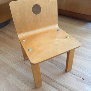 May include: A small, light-colored wooden chair with a rounded back and a circular cutout. The seat is rectangular, and the chair has four square legs. The chair is held together with silver-colored hardware.