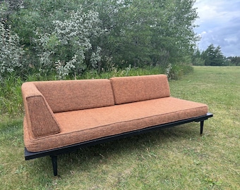 May include: A mid-century modern sofa with a burnt orange fabric and a black metal frame. The sofa has a low profile with a single armrest on the left side and two back cushions. The sofa is set on a grassy lawn with trees in the background.