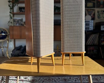 May include: A pair of vintage wooden speaker cabinets with a light brown wood finish and a gray fabric grille. The speakers are sitting on a light brown wooden table with four legs.