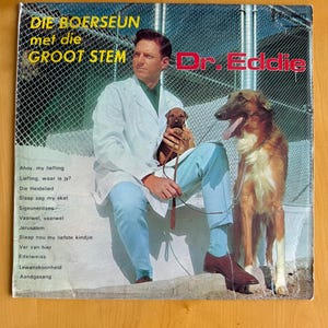 May include: Vintage album cover featuring a man in a white coat, seated with a small puppy and a large dog. The album title reads "DIE BOERSEUN met die GROOT STEM" with "Dr. Eddie" in red text. The cover has a blue and green background.