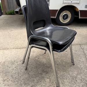 May include: A stack of two black molded plastic chairs with silver-toned metal legs. Each chair features a contoured seat and backrest with a horizontal cutout near the base of the back. The chairs have a smooth finish and a four-legged frame design.