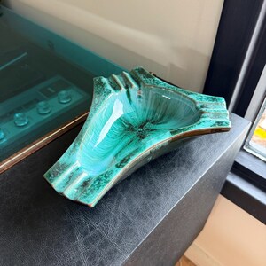 Peut inclure: Un cendrier vintage avec une forme unique et un glaçage turquoise vibrant. Le cendrier en céramique a un bord texturé et un bord marron foncé. L'intérieur présente un motif tourbillonnant de turquoise et de vert foncé. Une pièce élégante pour une décoration intérieure rétro.