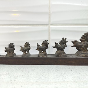 Può includere: Un set di sei piccole figurine di animali di colore bronzo disposte in fila su una base di legno scuro. Le figurine sono stilizzate e sembrano essere creature mitiche con ali e code.