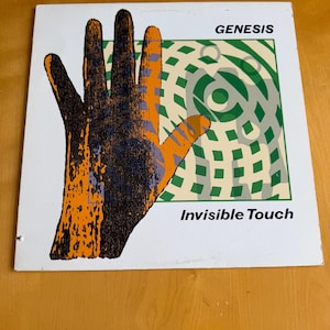 May include: Album cover for Genesis' "Invisible Touch." The cover features a stylized hand in orange and black over a green and white geometric design. The band name and album title are printed in black.