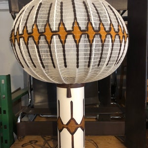 May include: A vintage table lamp with a white, globe-shaped shade featuring a woven pattern with brown and gold accents. The lamp has a white cylindrical base with brown geometric designs and a brown wooden base. The lamp is plugged in.