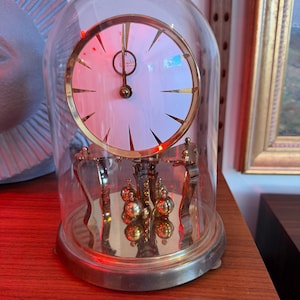 May include: A vintage brass and glass clock with a white face and gold hands. The clock is encased in a clear glass dome and sits on a wooden surface. The clock's pendulum features a series of brass spheres.