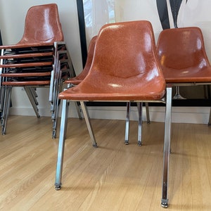 May include: A set of mid-century modern chairs with brown seats and chrome legs. Some chairs are stacked, while others are set up individually. The chairs have a classic, retro design, suitable for dining or accent seating.