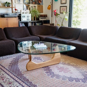 May include: A living room with a brown sectional sofa, a glass coffee table with a wooden base, and a patterned rug. The room has a mid-century modern style, with wooden cabinets and shelves.