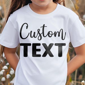 May include: A white short-sleeved t-shirt featuring the words Custom TEXT printed in black across the chest. The top line of text is in a cursive script font, while the bottom line is in a bold, sans-serif block font. The shirt has a crew neckline.