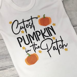 White short sleeve bodysuit on a wooden background embroidered with black text reading Cutest Pumpkin in the Patch and  orange pumpkins and little yellow stars.