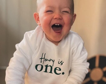 Puede incluir: Un niño pequeño con una camisa blanca de manga larga con el texto "Henry is one." en letras verdes, montando un juguete de madera con un mango blanco y ruedas grises.