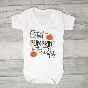 White short sleeve bodysuit on a wooden background embroidered with black text reading Cutest Pumpkin in the Patch and  orange pumpkins and little yellow stars.