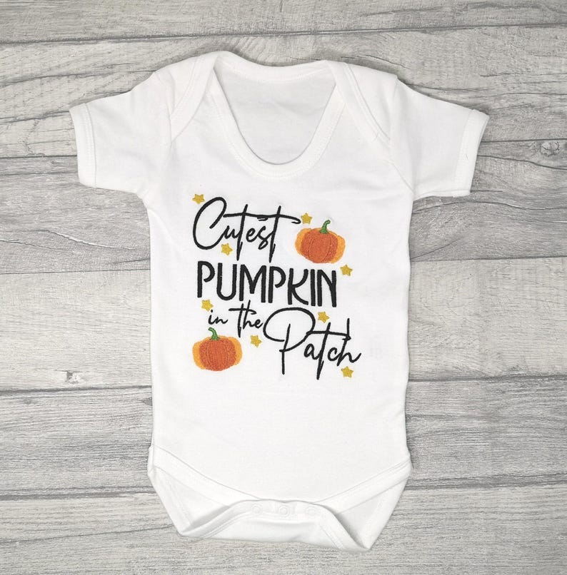 White short sleeve bodysuit on a wooden background embroidered with black text reading Cutest Pumpkin in the Patch and  orange pumpkins and little yellow stars.