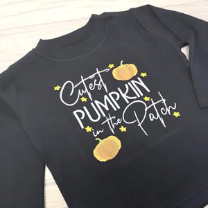 Long sleeve black t-shirt embroidered with black text reading Cutest Pumpkin in the Patch and  orange pumpkins and little yellow stars.