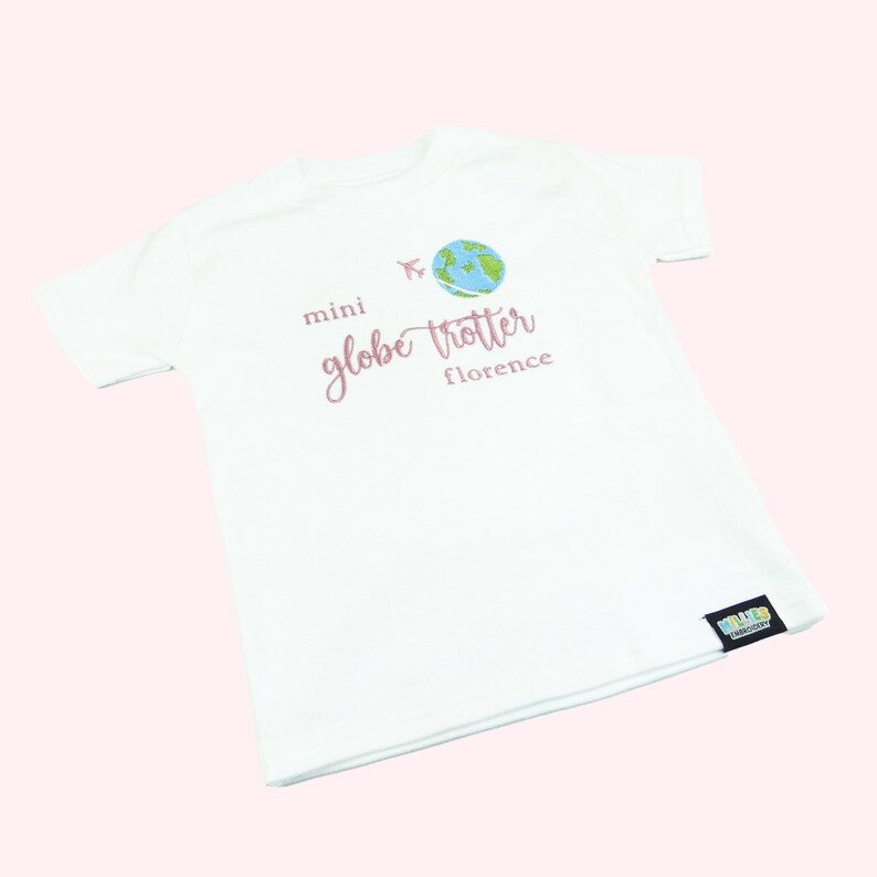 White tshirt embroidered with the phrase mini globe trotter  name and an embroidered earth with an airplane circling it.