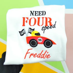 以下が含まれることがあります： 白地のTシャツに「NEED FOUR speed」の文字が黒と赤でプリントされています。赤いレーシングカーに黒い車輪と黄色のヘルメットが刺繍されています。名前の「Freddie」は赤で刺繍されています。