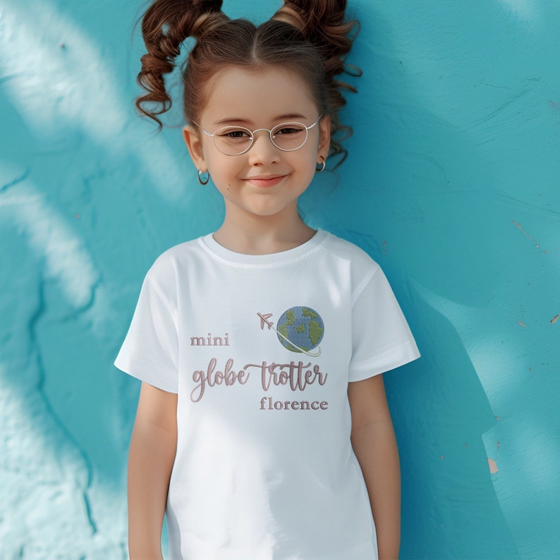 Young girl wearing a short sleeve white tshirt embroidered with the phrase mini globe trotter  name and an embroidered earth with an airplane circling it.