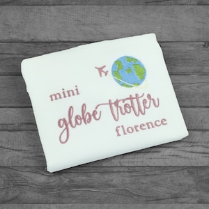 Folded white tshirt embroidered with the phrase mini globe trotter  name and an embroidered earth with an airplane circling it.