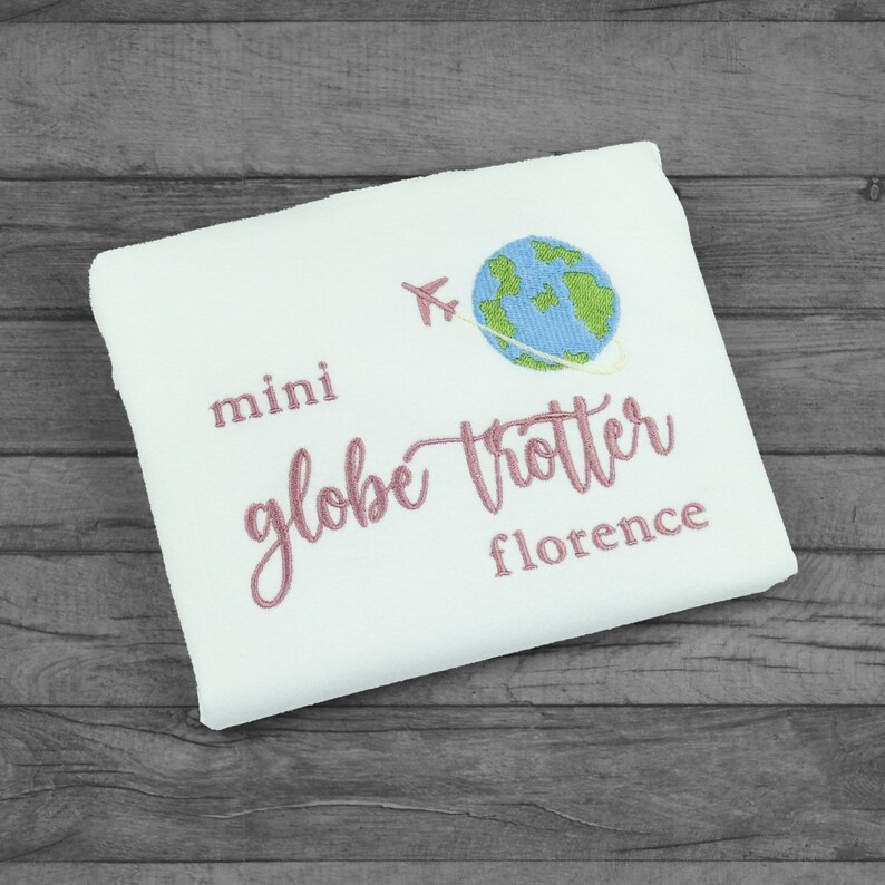 Folded white tshirt embroidered with the phrase mini globe trotter  name and an embroidered earth with an airplane circling it.