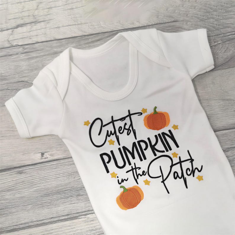 White short sleeve bodysuit on a wooden background embroidered with black text reading Cutest Pumpkin in the Patch and  orange pumpkins and little yellow stars.