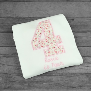 May include: White onesie with a pink floral number four and the text "Rosie is Four" embroidered on it.
