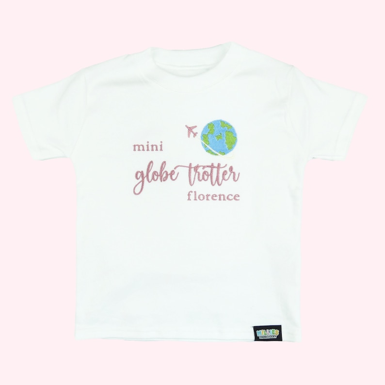 White tshirt embroidered with the phrase mini globe trotter  name and an embroidered earth with an airplane circling it.