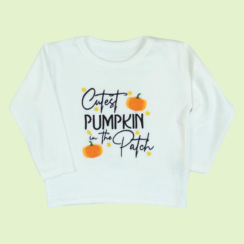 Long sleeve white t-shirt embroidered with black text reading Cutest Pumpkin in the Patch and  orange pumpkins and little yellow stars.