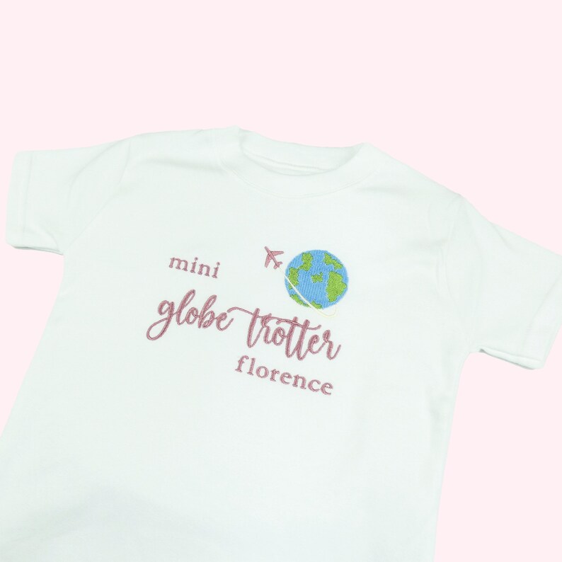White tshirt embroidered with the phrase mini globe trotter  name and an embroidered earth with an airplane circling it.