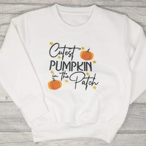 White jumper embroidered with black text reading Cutest Pumpkin in the Patch and  orange pumpkins and little yellow stars.