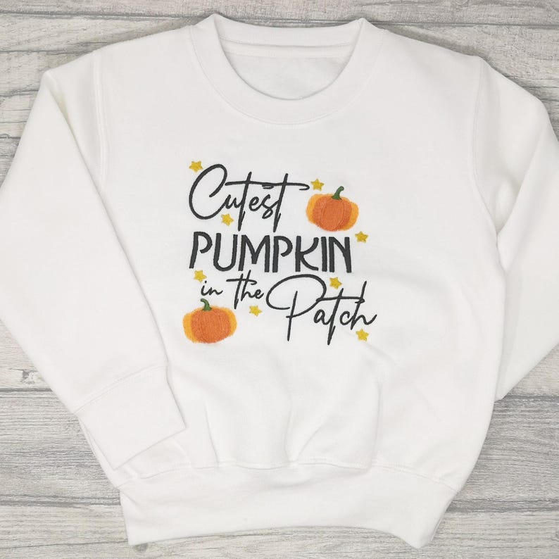 White jumper embroidered with black text reading Cutest Pumpkin in the Patch and  orange pumpkins and little yellow stars.