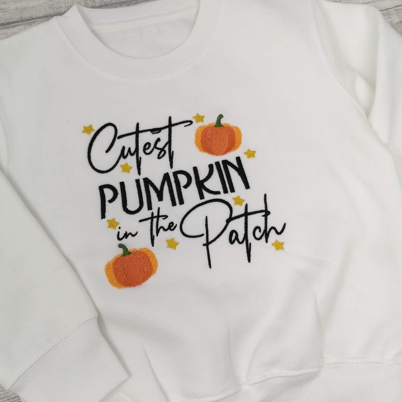 White jumper embroidered with black text reading Cutest Pumpkin in the Patch and  orange pumpkins and little yellow stars.