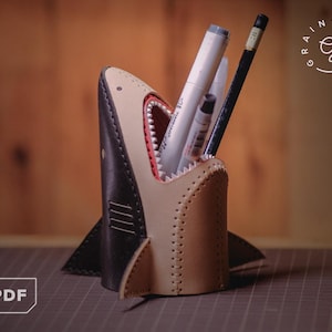 May include: A handmade shark-shaped pen holder made of leather. The pen holder is black and tan with a red interior, and it is filled with pens and pencils. The design includes fins and a stitched exterior. The logo "Rainy Days" is in the upper right corner.