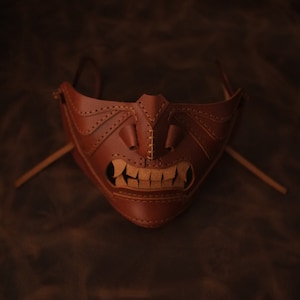 Pu&ograve; includere: Una maschera in pelle marrone con un design cucito che ricorda un viso demoniaco. La maschera ha una bocca larga con i denti e due piccoli occhi.