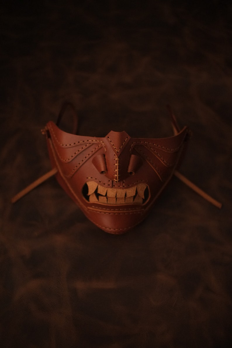 Pu&ograve; includere: Una maschera in pelle marrone con un design cucito che ricorda un viso demoniaco. La maschera ha una bocca larga con i denti e due piccoli occhi.