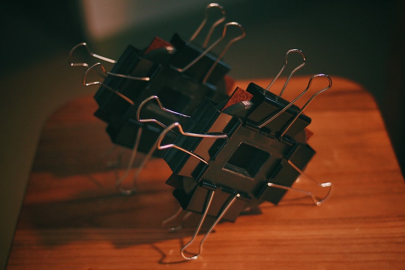 May include: Two black square frames with silver binder clips attached to them. The frames are stacked on top of each other and are sitting on a wooden surface.