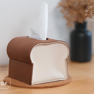 May include: A brown and cream-colored tissue box shaped like a slice of bread, with a white tissue sticking out of the top. The tissue box is sitting on a cork coaster. The background includes a black vase and dried flowers.