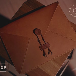 May include: A brown leather envelope-style laptop case with a button closure and a strap. The case is sitting on a wooden surface. The logo "Rainy Days" is visible in the upper right corner.