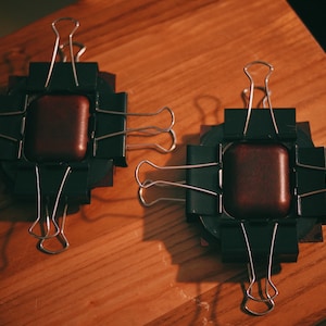 May include: Two brown leather squares are clamped together with black binder clips on a wooden surface. The clips are arranged in a cross pattern.