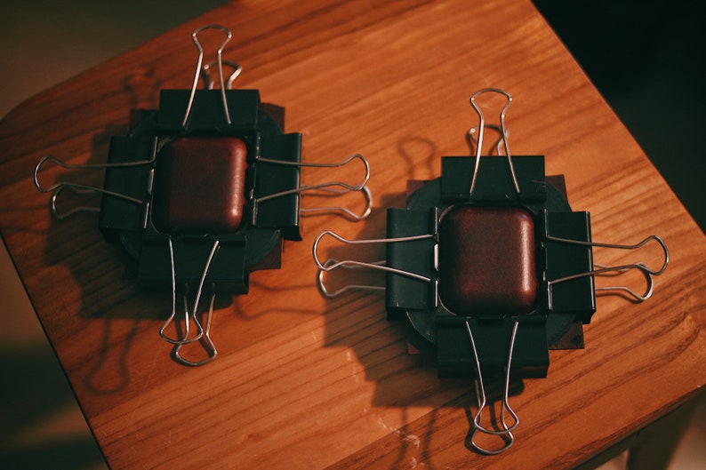 May include: Two brown leather squares are clamped together with black binder clips on a wooden surface. The clips are arranged in a cross pattern.