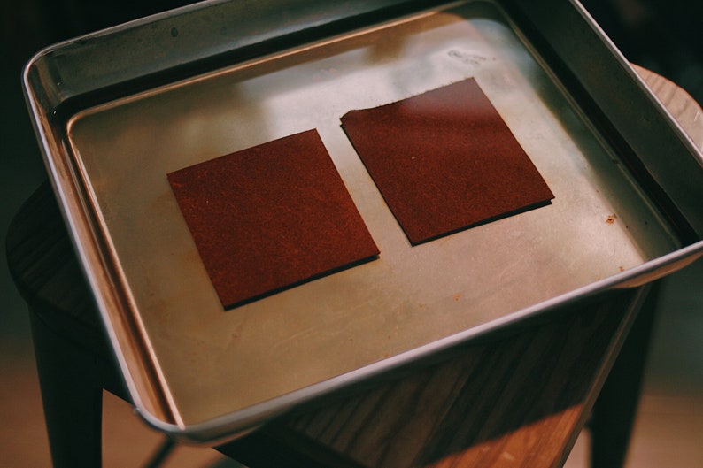 May include: Two brown leather squares submerged in a metal tray filled with water.