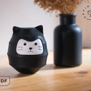 May include: A black leather cat-shaped decorative item with a white face and stitched details. The item has a rounded body and cat ears. A black vase and dried flowers are in the background. The text "GRAINY DAYS" is in a circle.