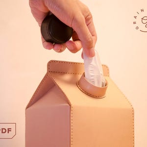 以下が含まれることがあります： 牛乳パックの形をしたベージュのレザー製ティッシュケース。縁にはステッチが施されています。上部の円形の開口部から白いティッシュが引き出されています。隅にはGrainydaysのロゴとPDFアイコンが表示されています。