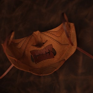 Pu&ograve; includere: Una maschera in pelle marrone con un design cucito e un'apertura per la bocca. La maschera ha due lunghi lacci in pelle per legare.